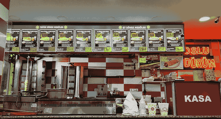 The counter of a döner shop: a lit menu board of sandwiches and price tags over two vertical spits and a griddle, red-and-white checkered tile behind, and a red register stand marked KASA.