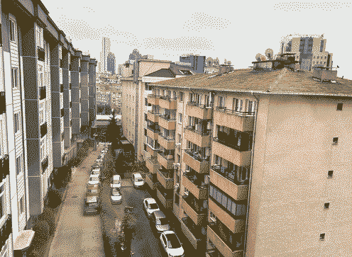 A narrow street seen from an upper window, cars parked nose to tail along it between pale apartment blocks with balconies and a tiled roof, a few towers in the distance.