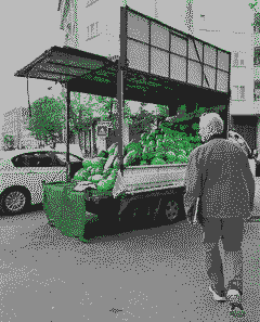 A man with white hair, in a gray jacket, walking past a small truck piled high with watermelons, parked beside a yellow taxi on an Istanbul street.